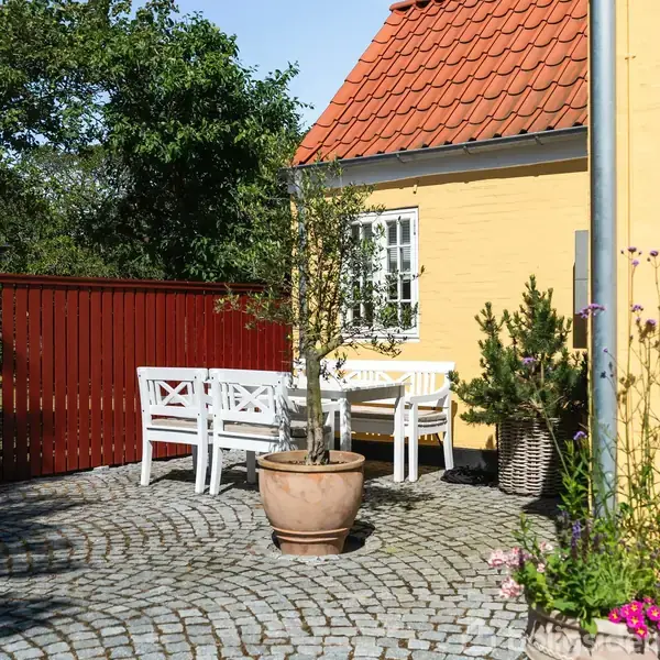 En hvid træbænk og bord står på en brostensbelagt terrasse ved en gul murstensvæg. Der er en krukke med et træ og en kurv med en busk.