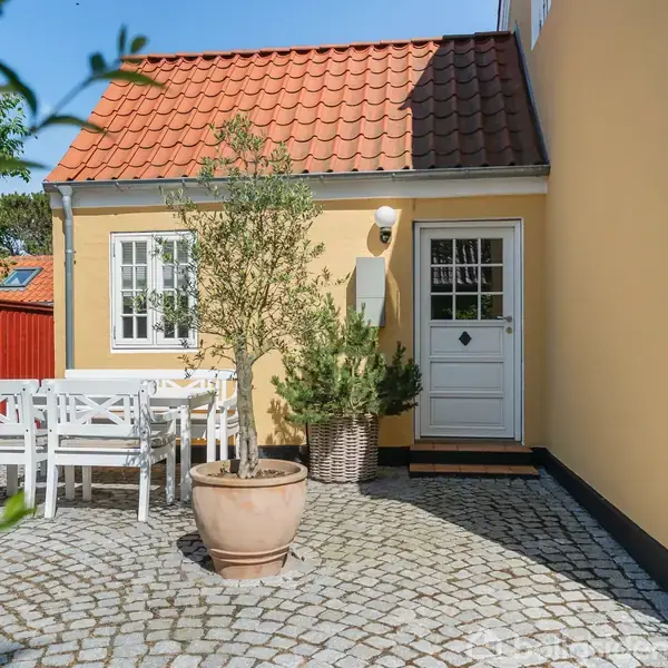 En gulmalet bygning med rødt tag, en hvid hoveddør, og vindue. I forgrunden står havemøbler og planter i krukker på en stenterrasse, omgivet af grøn beplantning.