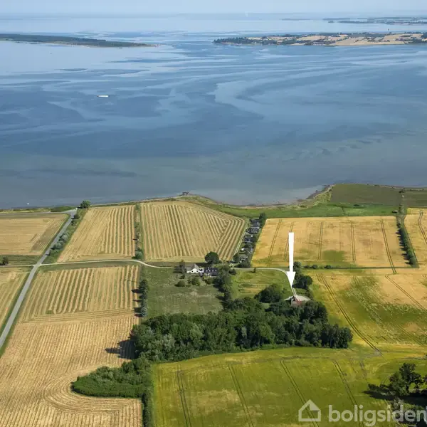 En gårdplads er omgivet af grønne træer og marker, med udsigt over en stor sø eller hav i baggrunden. En hvid pil peger mod gården.
