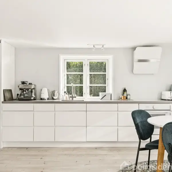 En grå garage med to trædøre står ved en grusvej. Den er omgivet af grønne træer og en græsplæne, der skaber en landlig atmosfære.