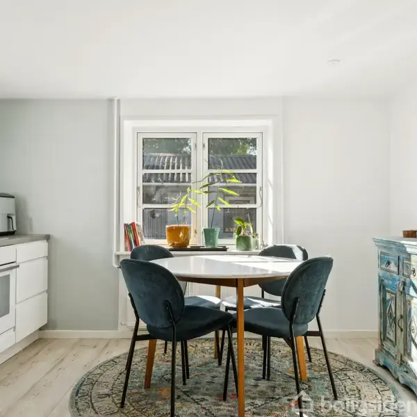 Et landligt hus med gråt tag og flere vinduer står omgivet af marker og træer. Et lille gårdhavemiljø med fliser og en stol er synlig foran huset.
