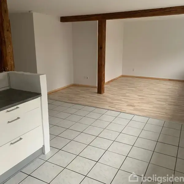 Køkken og stue i åben forbindelse. Køkkenet har hvide skabe og grå bordplade, mens stuen har lyst trægulv. Rummet har fritstående bjælker og hvide vægge.