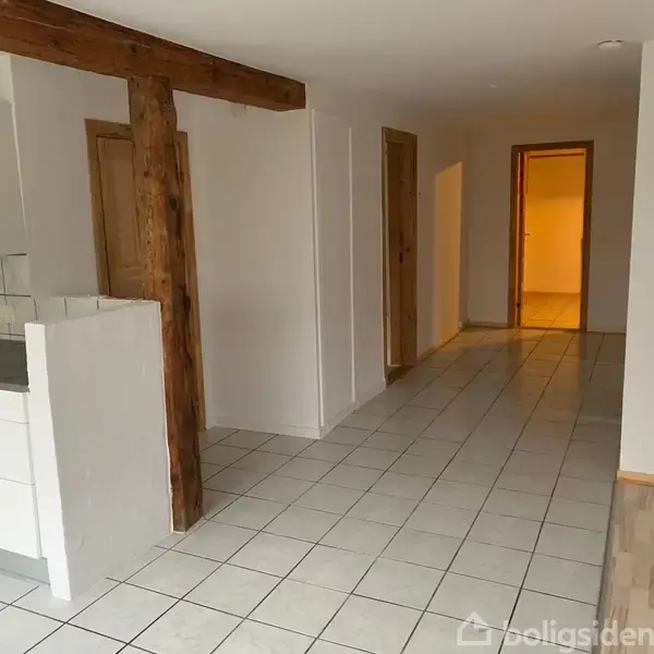 Køkkenområde med hvide skabe står stille ved siden af en fritliggende træbjælke. Flisebelagt gulv strækker sig mod en åben gang med flere døre.