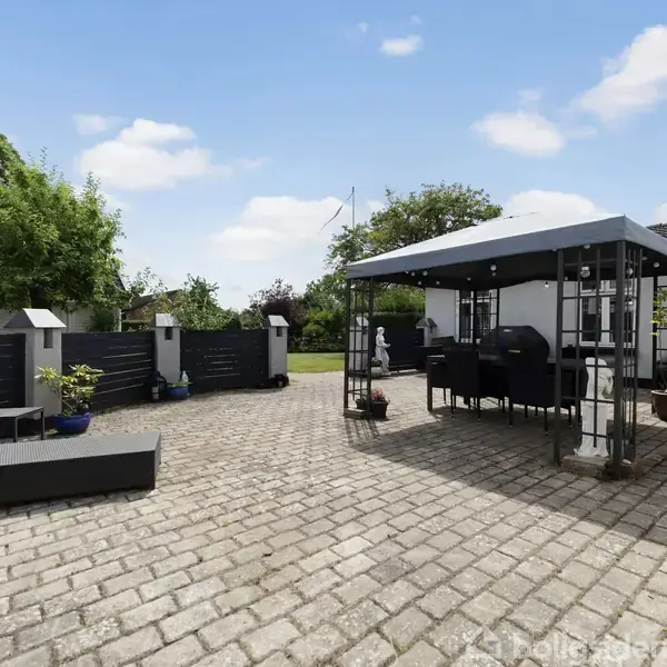 Terrasse med flisebelægning har en pavillon med havemøbler. Området er omgivet af sort hegn, planter i krukker og frodige træer i baggrunden under en klar blå himmel.