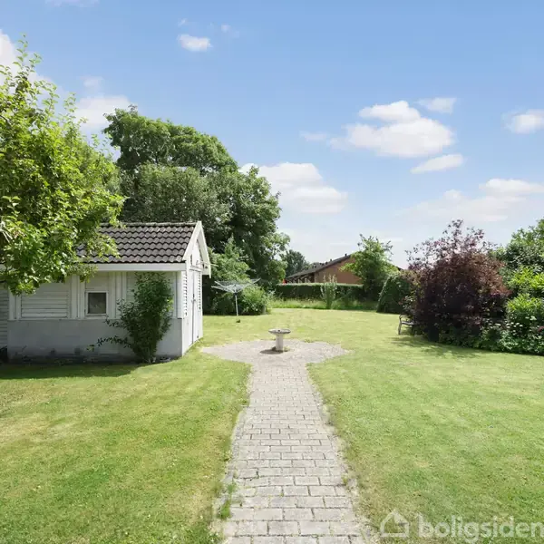 Lille hvidt hus står i en velplejet have. En flisebelagt sti fører forbi huset til en fuglebad, omkranset af frodig grønt og træer under en blå himmel med skyer.