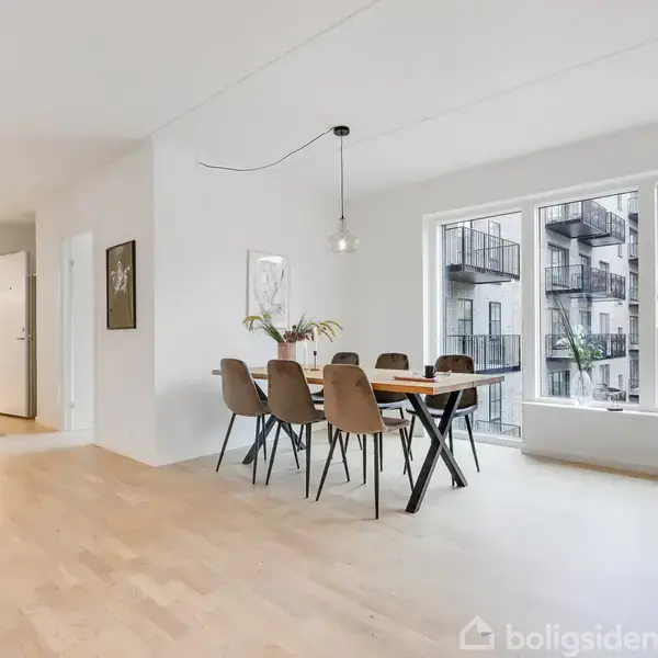 Spisebord med seks stole står i en lys, moderne lejlighed med trægulve. Store vinduer giver udsigt til en nærliggende bygning med altaner. En lampe hænger over bordet.