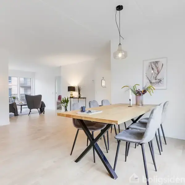 Bord i lys stue med seks stole, dekoreret med planter og lamper, placeret på trægulv nær sofagruppe og stor vinduesparti. En moderne og indbydende indretning.