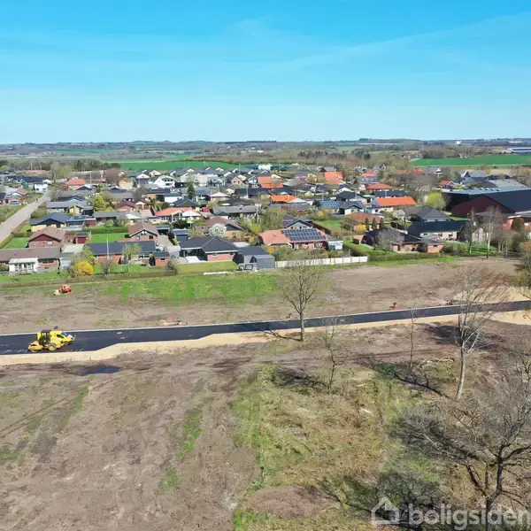 En vejmaskine arbejder på en nyanlagt vej i et område under udbygning. Omkringliggende huse og et grønt landskab ses under en klar blå himmel.