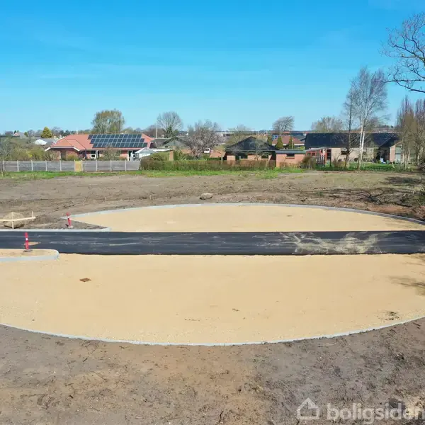 En cirkulær sandflade med en asfalteret vejstrækning i midten er omgivet af en åben jordplads. I baggrunden ses et boligområde med huse og træer.