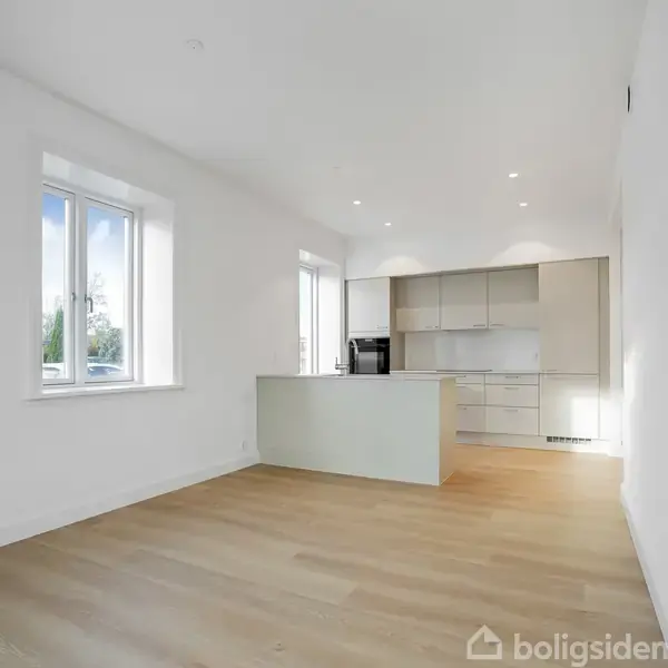 Et moderne køkken med hvide skabe og indbygget ovn står i et lyst rum med store vinduer og trægulv, der giver en rummelig atmosfære.