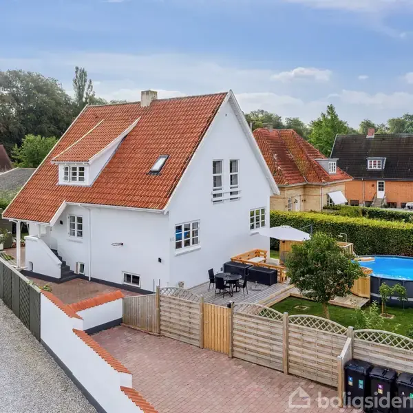 Et hvidt hus med rødt tegltag står ved en grusvej. I haven er der et rundt, blåt badebassin, terrasse med bord, og grønne omgivelser. Andre huse ses i baggrunden.