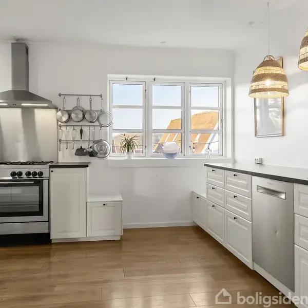 Et moderne køkken med en stor komfur og ovn til venstre, omgivet af hvide skabe. To flettede lamper hænger over en sort bordplade. Et vindue viser udsigt til tage.