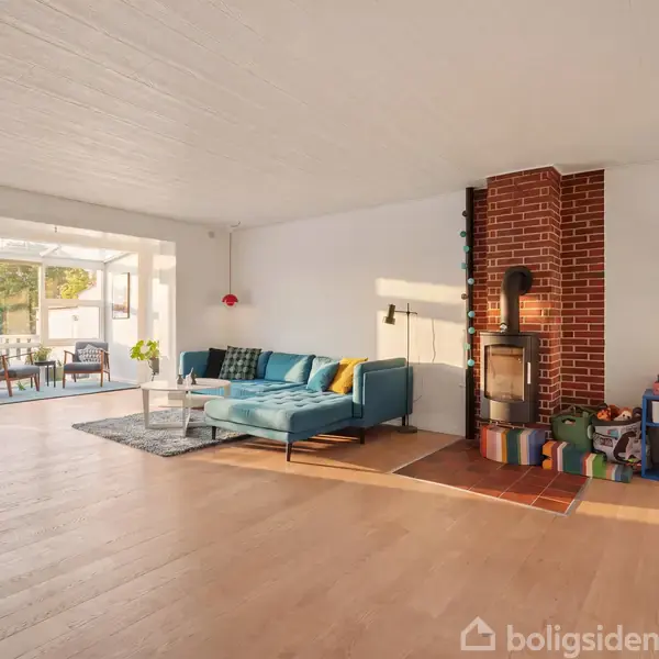 En stue med blå hjørnesofa, sofabord og tæppe; brændeovn indbygget i murstensvæg. Et legeområde er til højre med farverige hylder. Store vinduer og terrasse i baggrunden.