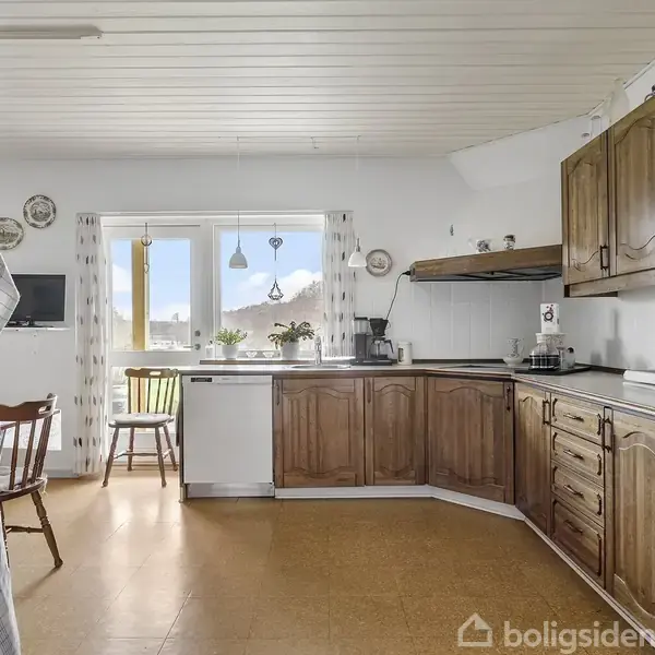 Køkken med træskabe står stille i et lyst rum. Et bord og stol ses ved vinduer. Køkkenredskaber er synlige, og natur ses udenfor vinduet. Ingen tekst til stede.