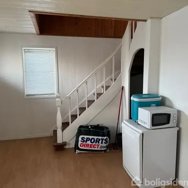 En trappe fører opad ved siden af en hvid væg. Under trappen står en taske med "Sports Direct.com". Ved siden af er et mini-køleskab med en mikroovn og en køletaske ovenpå.