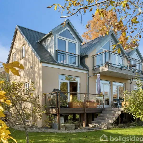 Hus med to etager har store vinduer og altaner, beliggende i en have med grønne græsplæner og træer med efterårsfarver.