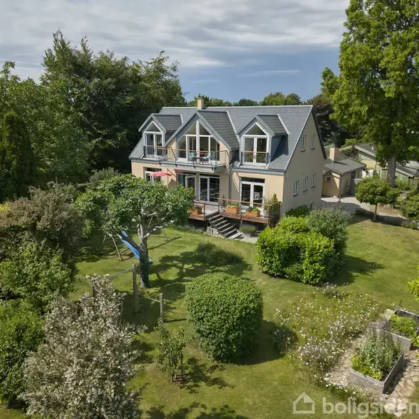 Et stort hus med grå tagsten og balkon står i en frodig have med tætte buske og træer. En blå hængekøje hænger fra et træ i forgrunden.