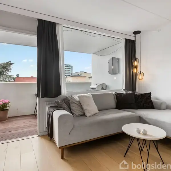 Grå sofa med puder står i en stue med hvide vægge. Ved siden af er en altan med træterrasse og blomsterpotte. Store vinduer lukker lys ind.