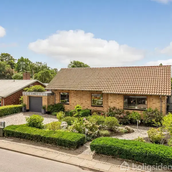 Et rødt murstenshus med tegltag står i et velholdt haveområde omgivet af grønne buske og træer. En tilknyttet garage er synlig, beliggende i et villakvarter.