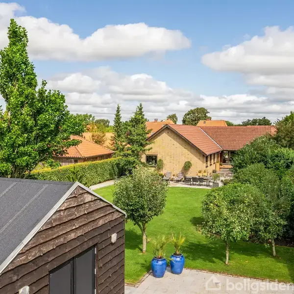 Et træhus står stille i en frodig have med grønne græsplæner og træer. I baggrunden ses et gult hus med rødt tegltag under en blå himmel med hvide skyer.