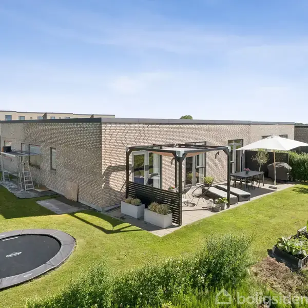 En moderne enfamiliehust består af en murstensbygning med fladt tag. Haven indeholder en nedgravet trampolin, terrasse med pergola og havesæt. Omkring ejendommen ses græsplæner og beplantning.