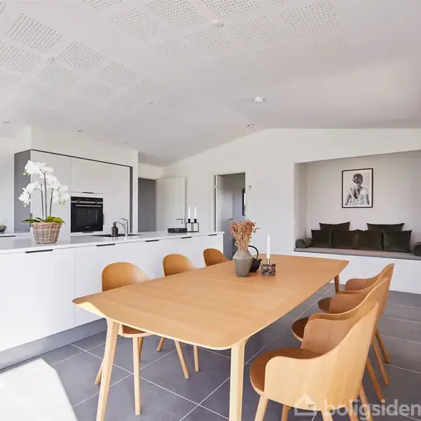 Et moderne køkken-alrum med en lys trælignende spisebord omgivet af seks stole, placeret på grå fliser. Et hvidt køkken med sort emhætte og hvidevarer i baggrunden.