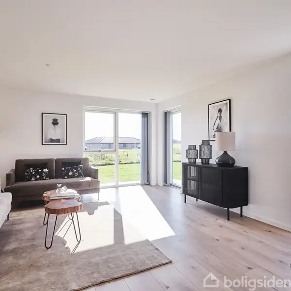 En lys stue med to sofaer og et sofabord. Lampe og billeder hænger på væggen. Stort vindue åbner op til en grøn have.