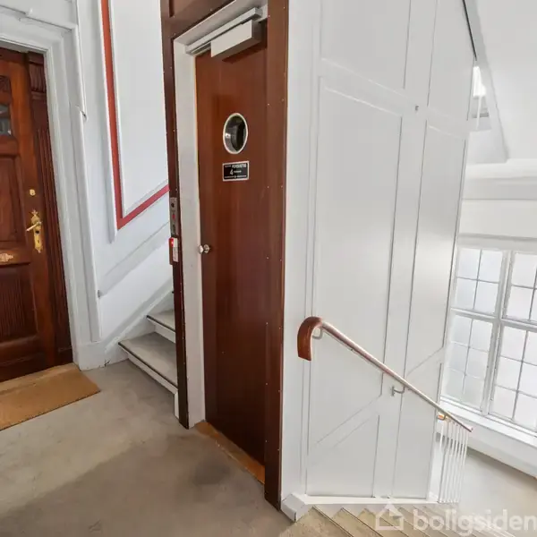 Dør med dekorativ krans står ved siden af en elevator med skiltet "Elevator ikke i drift". Trappe med gelænder i et lyst trappeopgangsmiljø.