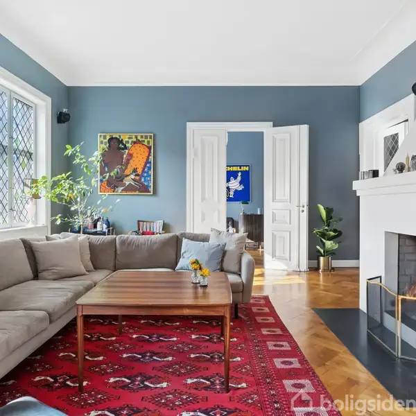 Stor beige sofa står i stue, omgivet af bogreoler og planter. Pejs med ild til højre. Rød mønstret tæppe dækker gulvet. Lys kommer ind fra et vindue. Veggpynt og åben dør i baggrunden.