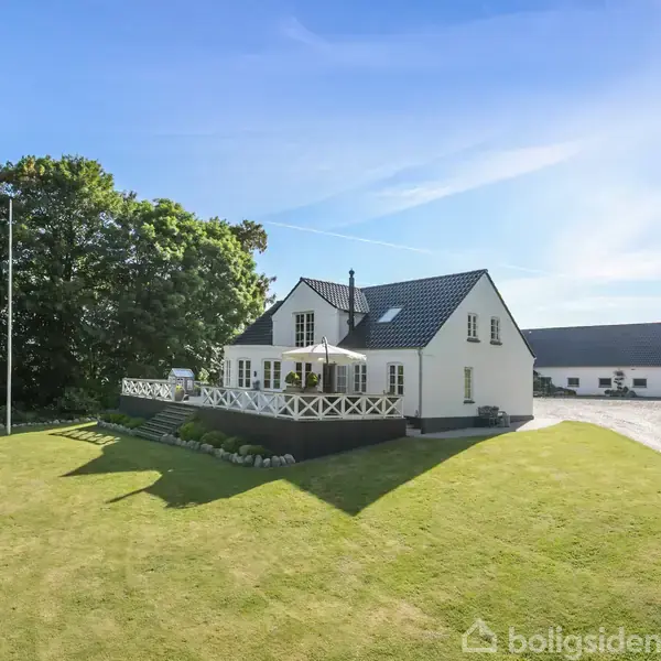 Et hvidt hus med sort tag står i en velholdt have med grøn græsplæne. Der er en terrasse med parasol og møbler. Omgivet af træer og en lang indkørsel.