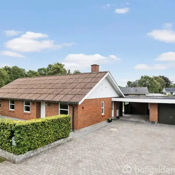 Et rødt murstenshus står stille i en veltrimmet have med en brolagt indkørsel, omgivet af grønne buske. Til højre ses en carport og flere træer i baggrunden.