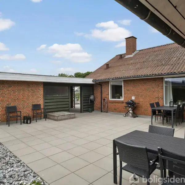 Rødt murstenshus med terrasse; borde og stole står på fliserne; en lille pejs ses i havebedet. Skyfri himmel og græsplæne omkranser terrassen.