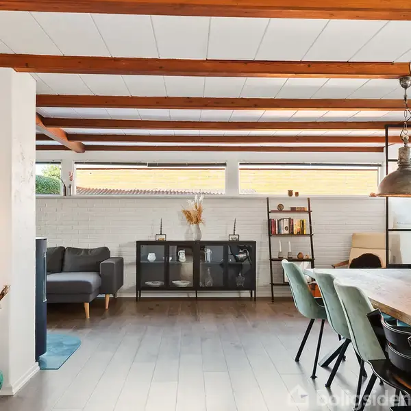 Stue med spisebord og stole, oplyst af hængelys. Sofa og skab mod hvidmalet murstensvæg. Træbjælker i loftet skaber et hyggeligt, moderne rum. Ingen tekst at bemærke.