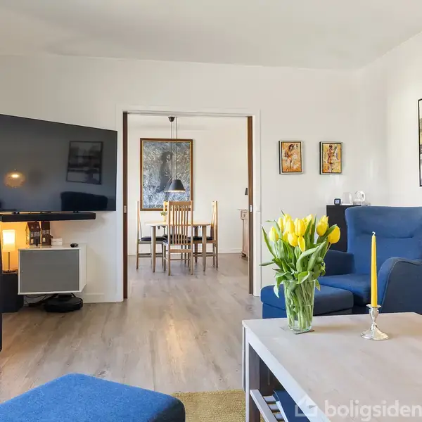 Stue med blå lænestol og sofabord med gule blomster og lys. Fladskærms-tv på væggen. Bagved ses spisebord i en tilstødende spisestue. Væggene er dekoreret med kunst.