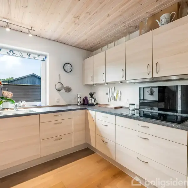 Et lyst køkken med træloft og -gulv. Hvide skabe med rustfri stållåger, sorte bordplader og moderne apparater. Et vindue med udsigt til en have med en enkelt plante ved vasken.