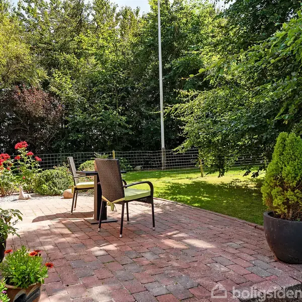 Terrassemøbler står på en stenbelagt patio omgivet af grønne planter og blomster i krukker. I baggrunden ses en frodig have med græsplæne og tætte træer.
