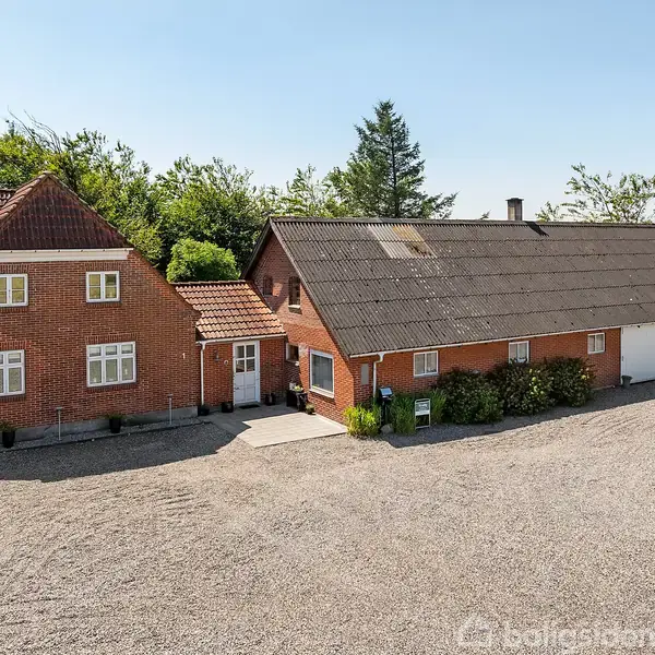 Rødt murstenshus med mørkt tag og flere døre står på en grusbelagt gårdsplads. Bag huset er der grønne buske og træer, der strækker sig mod en blå himmel.