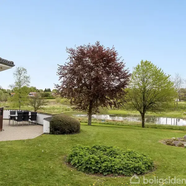 Et moderne hus med terrasse og havemøbler står ved en grøn have. En stor, rødbladet træ og en mindre, grøn træ ses nær en sø i baggrunden under en klar, blå himmel.
