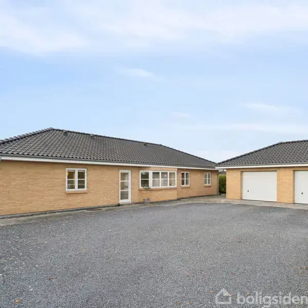 Hus i gule mursten står stille, omgivet af stor grusplads og garagen med to porte ved siden af. Himlen er blå med hvide skyer.