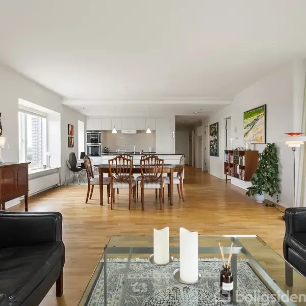 Sorte lænestole står foran et glas sofabord med stearinlys og pyntegenstande i en lys stue. I baggrunden ses et spisebord med træstole og et moderne køkken.