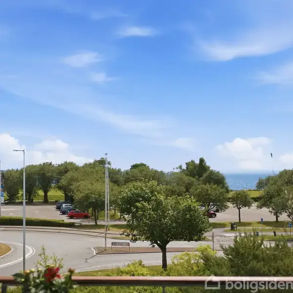 Rundkørsel omgivet af grønt løv og træer. I baggrunden skimtes havet under en blå, let skyet himmel. Biler er parkeret på venstre side af vejen.