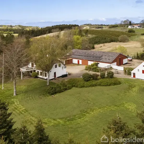 En gård med hvidmalede bygninger og røde tage står omgivet af grønne marker og træer. Flere bygninger er synlige, herunder en stor lade. Landskabet er landligt og frodigt.