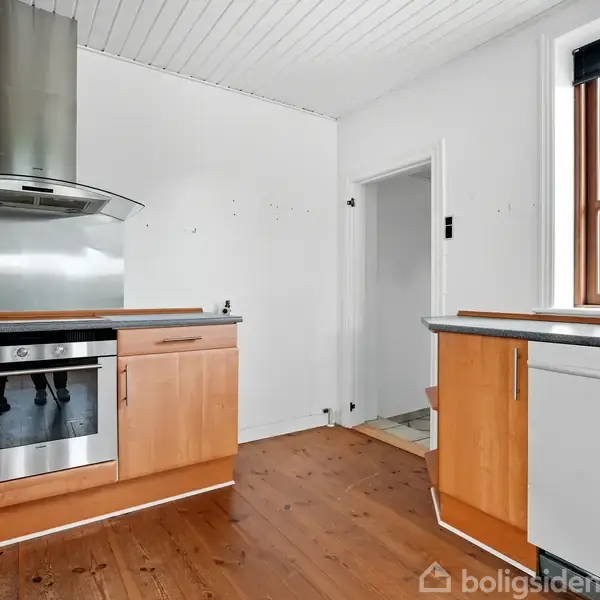 Et køkken med træskabslåger og indbygget ovn. En emhætte er monteret over komfuret. Rummet har lyse vægge og trægulv, med et vindue til højre, der giver naturligt lys.