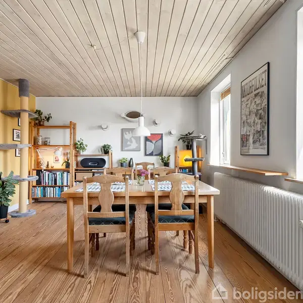 Spisebord med seks stole er dækket op i en lys spisestue. Hylder med bøger og planter omkranser rummet, og store vinduer trækker naturligt lys ind.