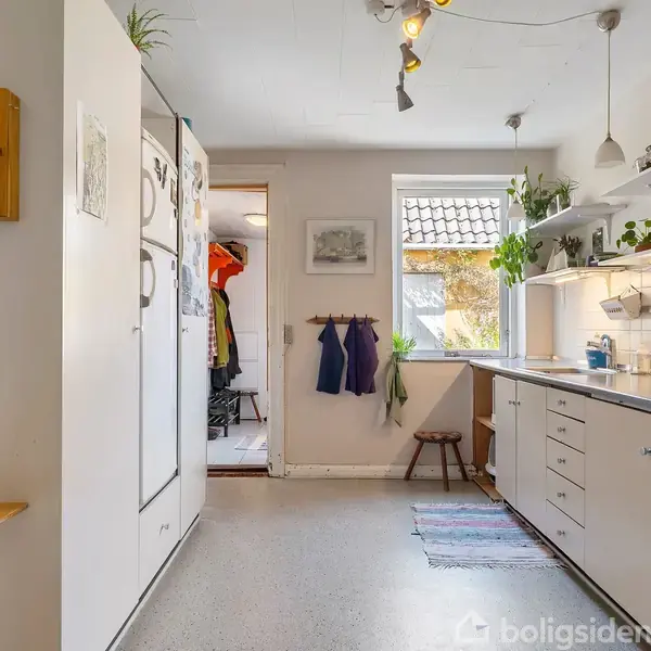 Køkken med hvide skabe og bordplader, hvor planter dekorativt står på hylder. En kedel står på komfuret. Et stort vindue giver lys, med en døråbning mod gangen.