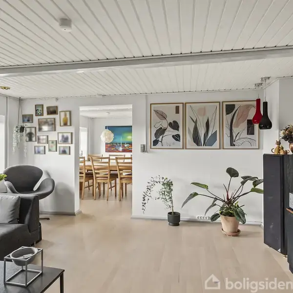 Stue med sofa og reol under vægkunst. Plakater hænger over planter ved indgangen til et spisestueområde med træstole. Væggene er hvide med mange dekorationer.