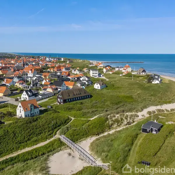 By består af farverige huse, ligger langs kysten med en bro i forgrunden. Strand og hav skaber en fredfyldt atmosfære under en klar blå himmel.