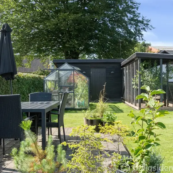 Et havebord med stole står på en terrasse ved siden af en lille have med et drivhus og et skur i baggrunden. Omgivelserne er grønne med buske og træer.