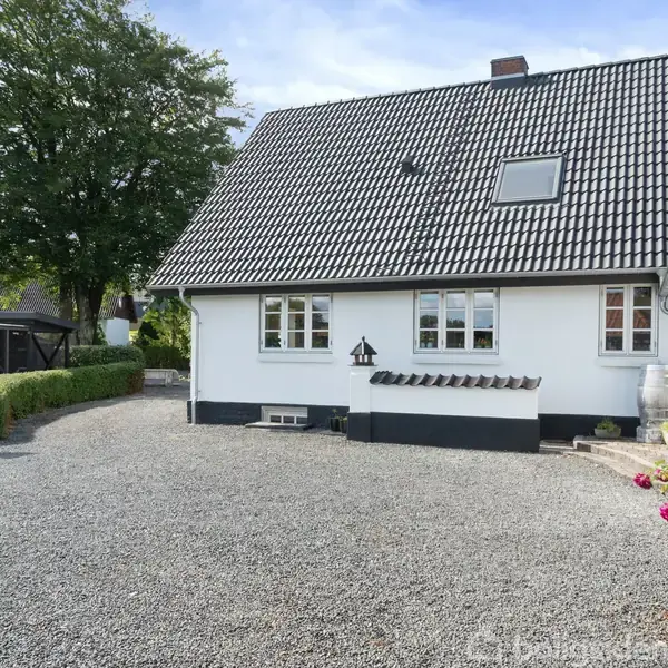 Brændeovn står tændt i hjørnet. Lysegråt opholdsrum med trægulv og træloft. Komfortable sofaer og stole omkring et sofabord. Vinduer og pyntegenstande skaber en hyggelig atmosfære.