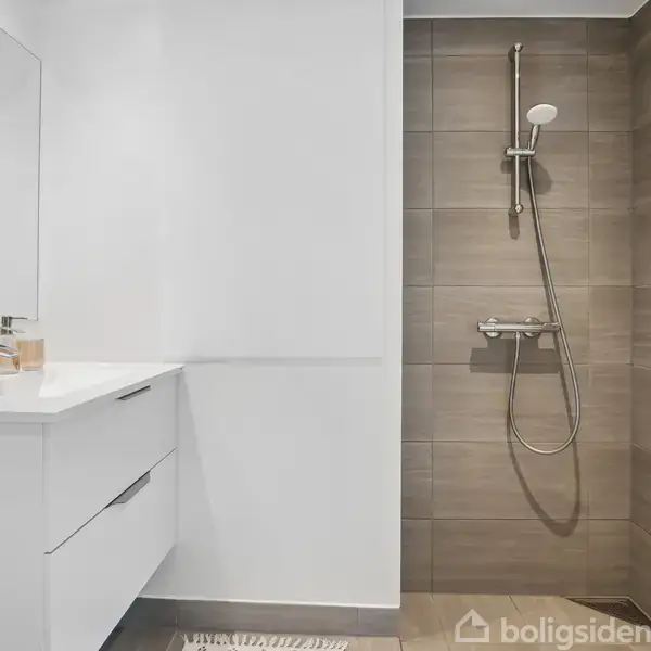 En moderne bruser hænger på en væg med beige fliser, ved siden af et hvidt badeværelsesmøbel med vask og spejl. Miljøet er minimalistisk og lyst.